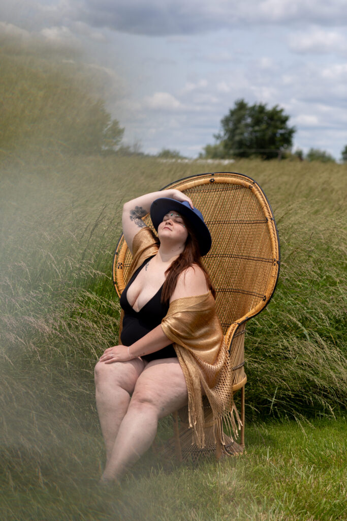 Woman sitting on a peacock chair in a black bodysuit and hat, surrounded by tall grass, showcasing elegance and empowerment at golden hour. Step Into the Sun: Outdoor Boudoir Photography in York, PA