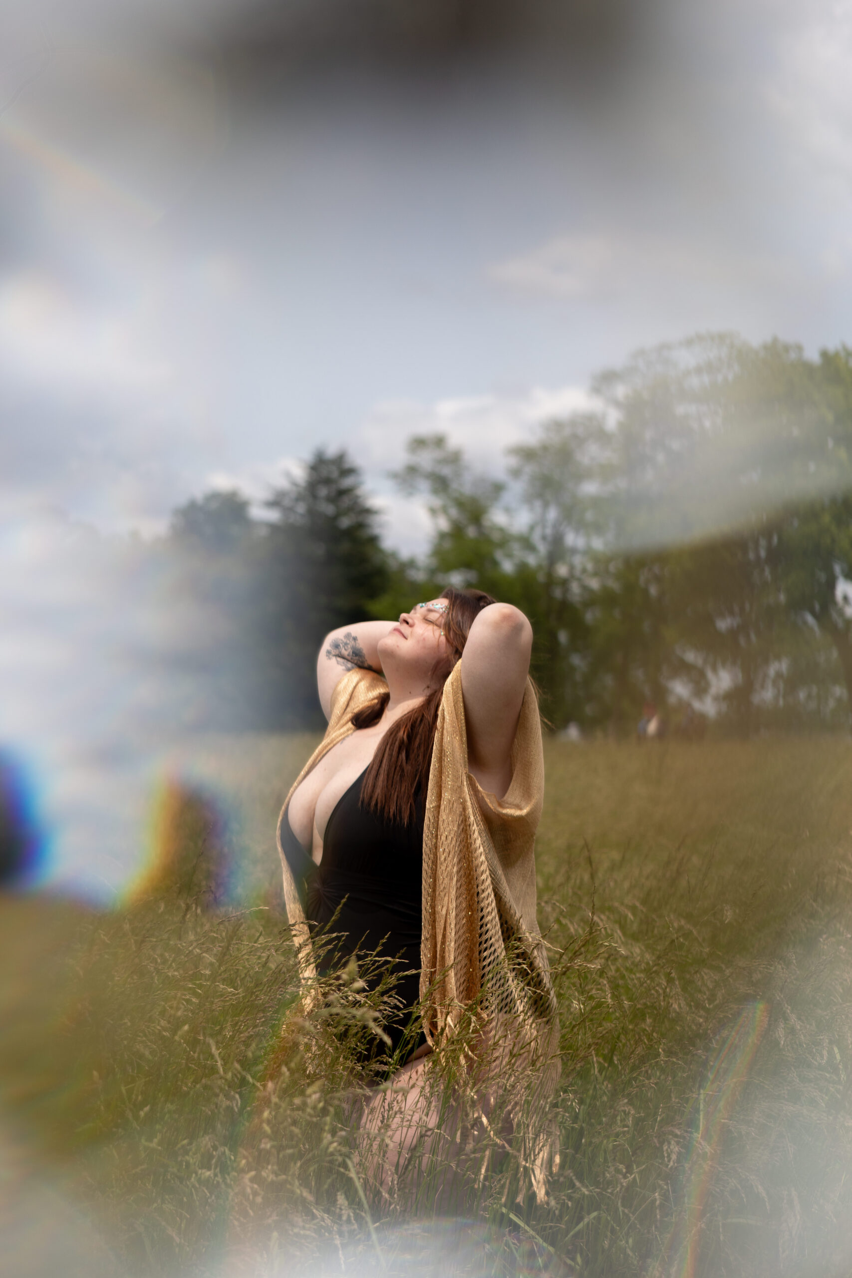 Woman standing in tall grass during outdoor boudoir session, wearing gold shawl and black bodysuit, bathed in soft golden sunlight.