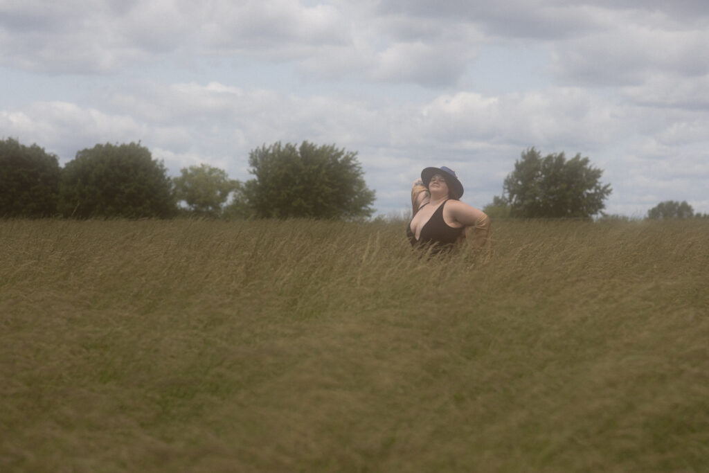 Woman in wide-brimmed hat standing in tall grass, celebrating body confidence during an empowering outdoor boudoir shoot.